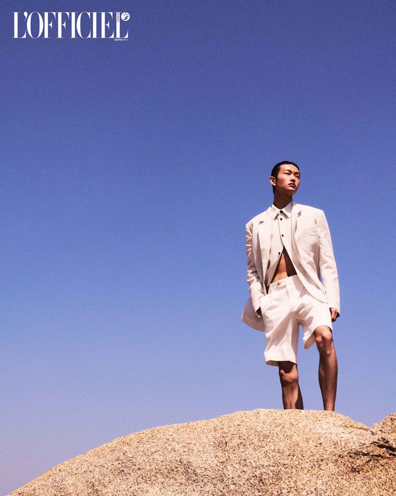 A man in an unbuttoned linen suit and shorts stands on a sandy dune against a clear blue sky in a fashion editorial shot.