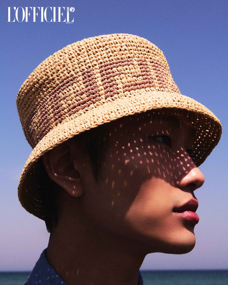 Profile portrait of a young adult male wearing a woven bucket hat outdoors against a blue sky and ocean background on a sunny day.