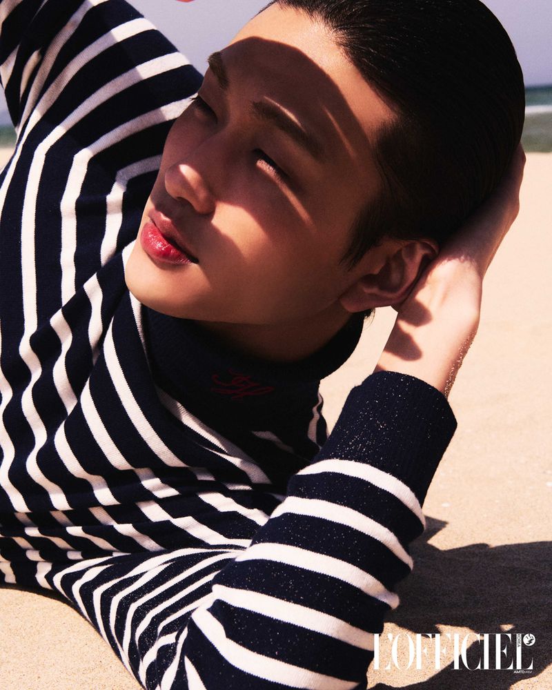 A fashionable Asian male model with dark hair lies on a tan beach in a striped shirt, posing for an outdoor fashion editorial shoot.