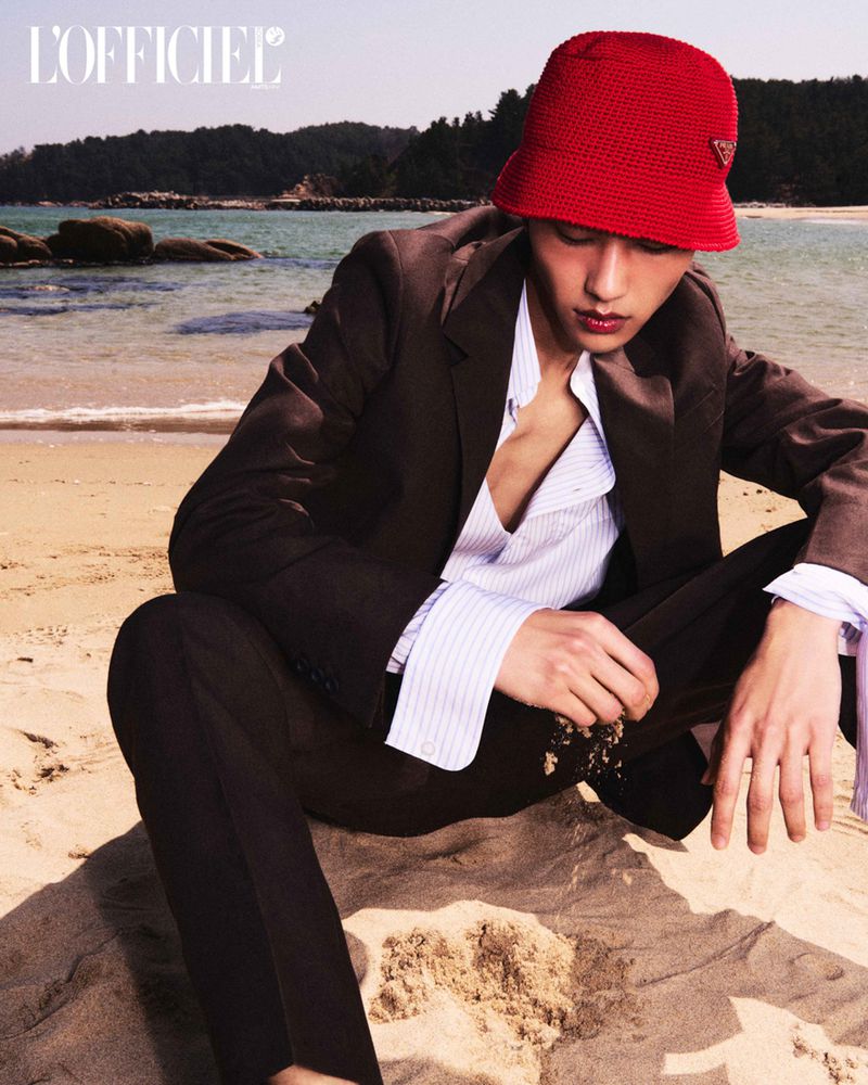Man in a brown suit sitting on the beach, wearing a red knit hat. The background features a beach with gentle waves and rocky outcroppings.