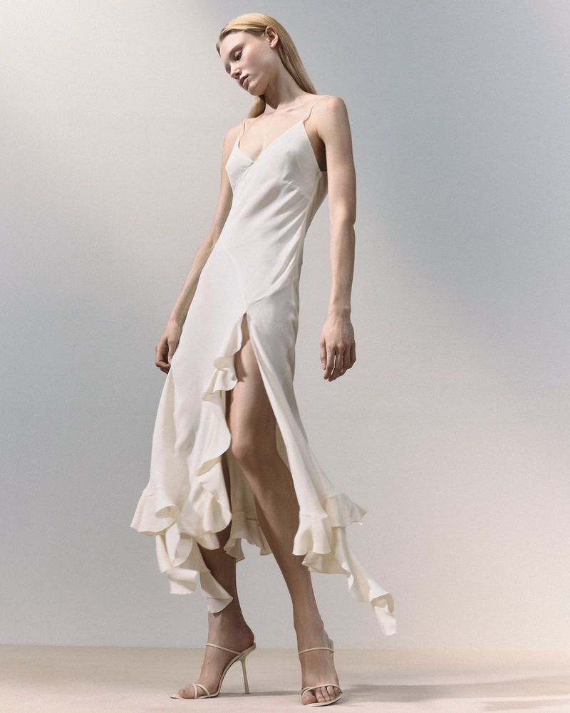 A blonde adult woman poses in a full-length shot wearing a spaghetti-strap dress with ruffled accents and high-heeled strappy shoes on a gradient background.