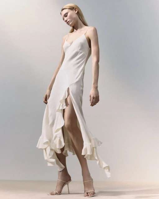 A blonde adult woman poses in a full-length shot wearing a spaghetti-strap dress with ruffled accents and high-heeled strappy shoes on a gradient background.