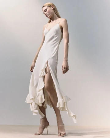 A blonde adult woman poses in a full-length shot wearing a spaghetti-strap dress with ruffled accents and high-heeled strappy shoes on a gradient background.