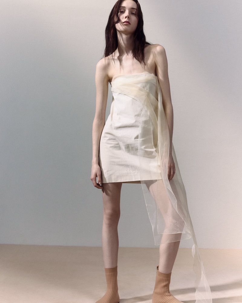 Elegant woman in a strapless mini dress poses in a light studio with soft, neutral tones and subtle shadows, showcasing fashion.