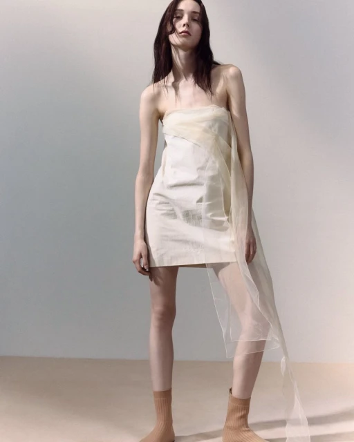 Elegant woman in a strapless mini dress poses in a light studio with soft, neutral tones and subtle shadows, showcasing fashion.
