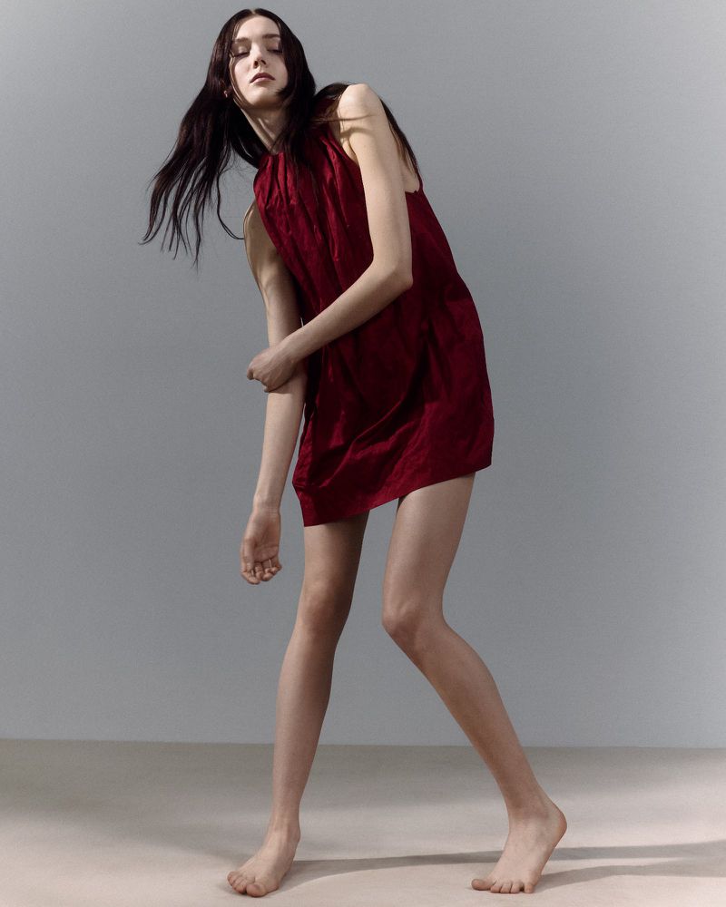 Full shot of a slender young woman with long dark hair wearing a dark red dress, posing barefoot in a neutral studio setting.