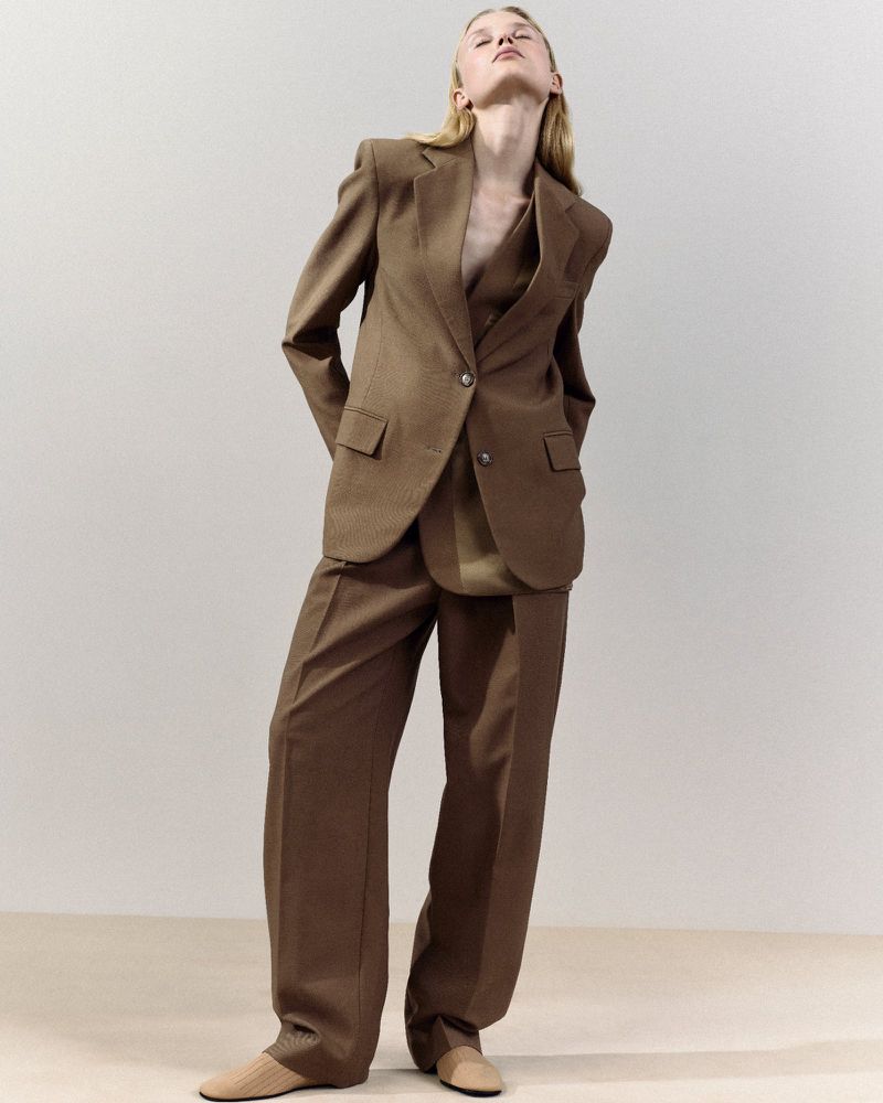 Woman in brown suit stands indoors, head tilted back, in a portrait displaying high-waisted pants and a matching blazer.