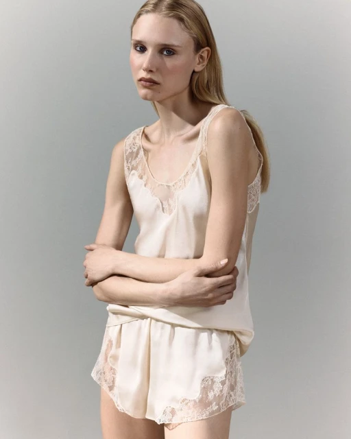 A blonde woman is wearing a light-colored lingerie set with lace trim in a studio shot. She is facing the camera with her arms crossed.