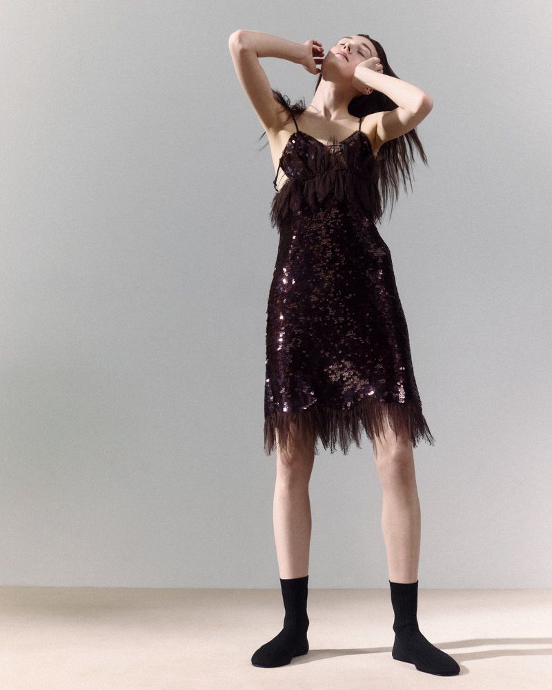 A slender woman in a shimmering, fringed slip dress poses in a photo studio, arms raised, wearing black socks and minimal makeup.