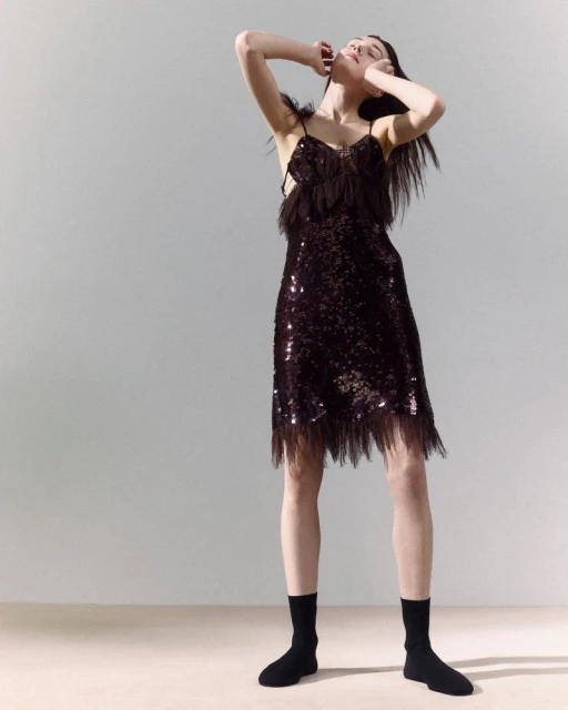 A slender woman in a shimmering, fringed slip dress poses in a photo studio, arms raised, wearing black socks and minimal makeup.