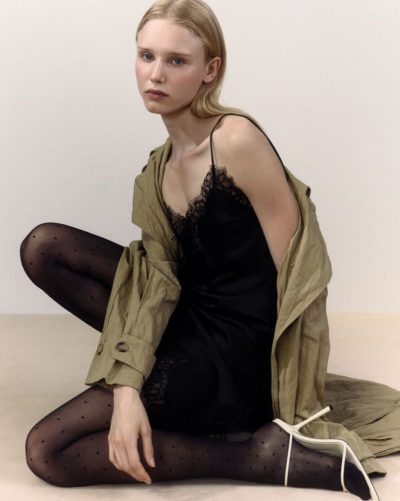 A fair-skinned model poses in a black, lace-trimmed slip dress, sheer polka dot tights, and a neutral trench coat against an off-white backdrop.
