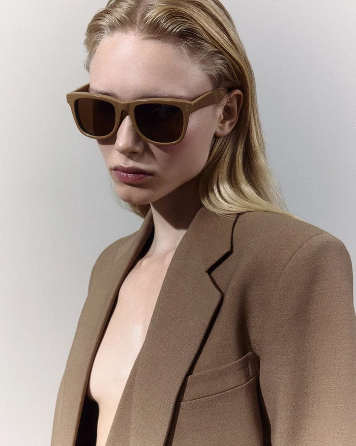 Model with blonde hair wearing oversized brown sunglasses and a tailored tan blazer against a light neutral background