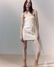 Model wearing a strapless off-white dress with sheer overlay and beige boots standing against a neutral background