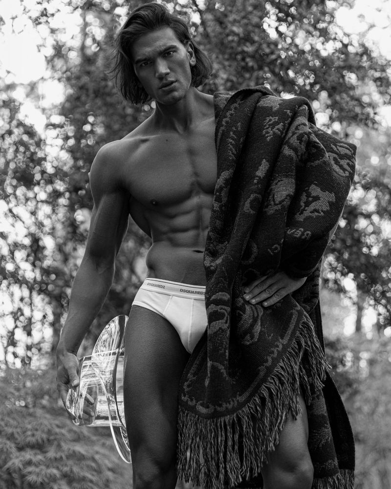 Monochrome portrait of a muscular man with long hair, wearing underwear and holding a shawl, posing outdoors with trees in background.