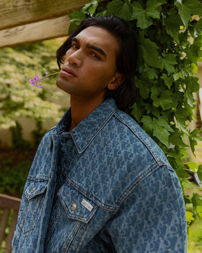 Portrait of a dark-haired male model wearing a patterned blue denim jacket, holding a purple flower in his mouth, leaning against ivy.