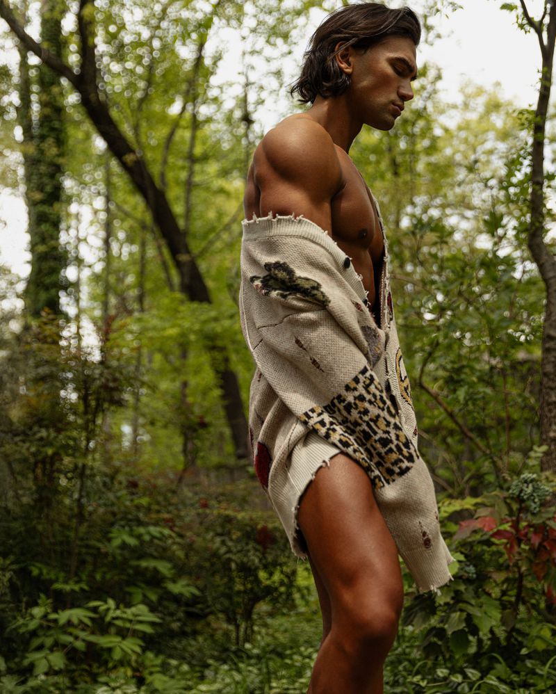 A shirtless man with long hair and a patterned sweater stands in a forest with greenery in the background in a medium shot.
