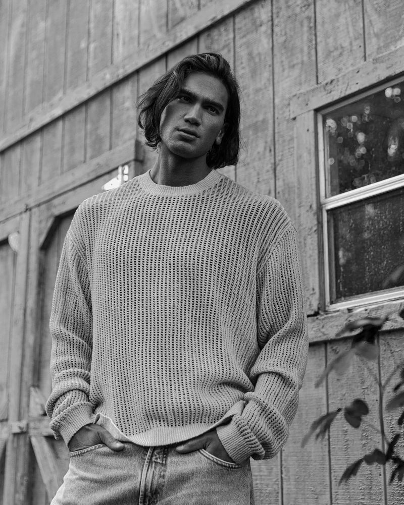 Monochrome portrait of a man in a knit sweater and jeans posing outdoors against a weathered wood building with a window.