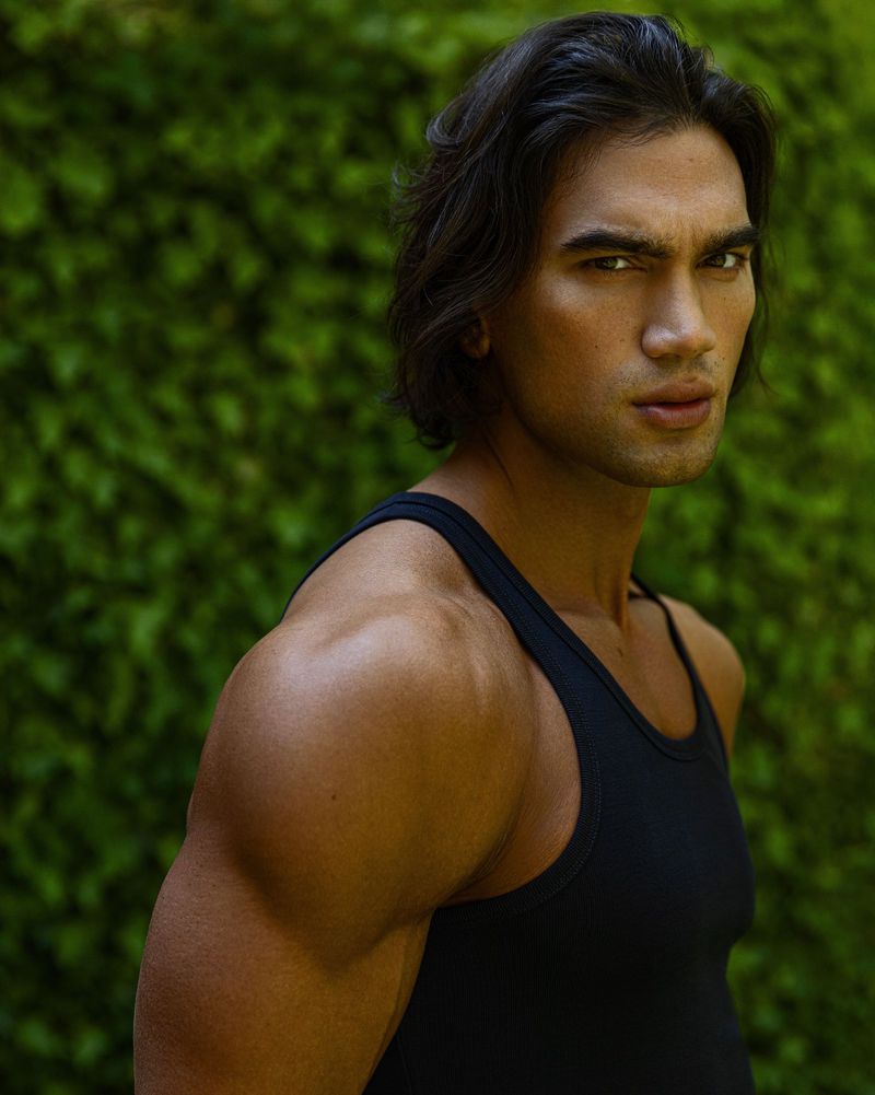 Handsome adult male with long dark hair poses confidently in a sleeveless shirt, showcasing muscular build, against a green foliage backdrop.