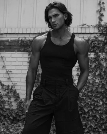 Monochrome portrait of a muscular man in a tank top and wide-legged pants, posing in front of a brick wall covered in ivy.