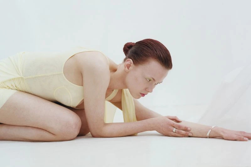 Woman in a light yellow dress kneels on a white surface with her arms extended, showcasing jewelry against a plain background.