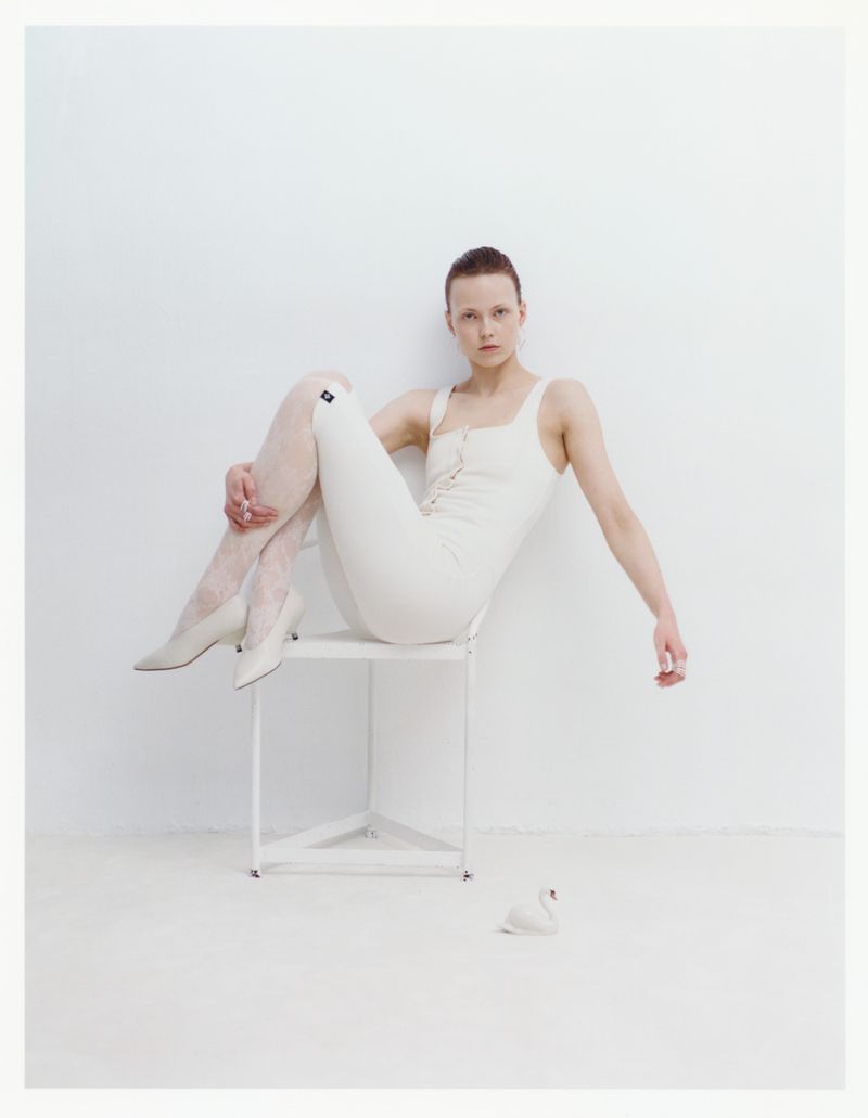Young woman in white tights and bodysuit sitting on a white chair in a white room with swan figurine on the floor. Fashion studio shot.