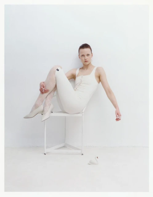 Young woman in white tights and bodysuit sitting on a white chair in a white room with swan figurine on the floor. Fashion studio shot.