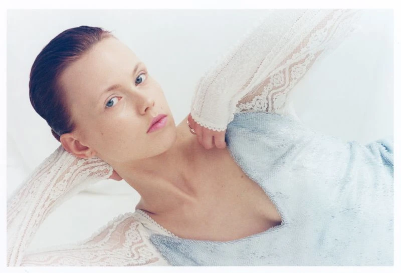 A fair-skinned woman lies against a white background wearing a light blue dress and a white lace top, gazing directly at the camera.