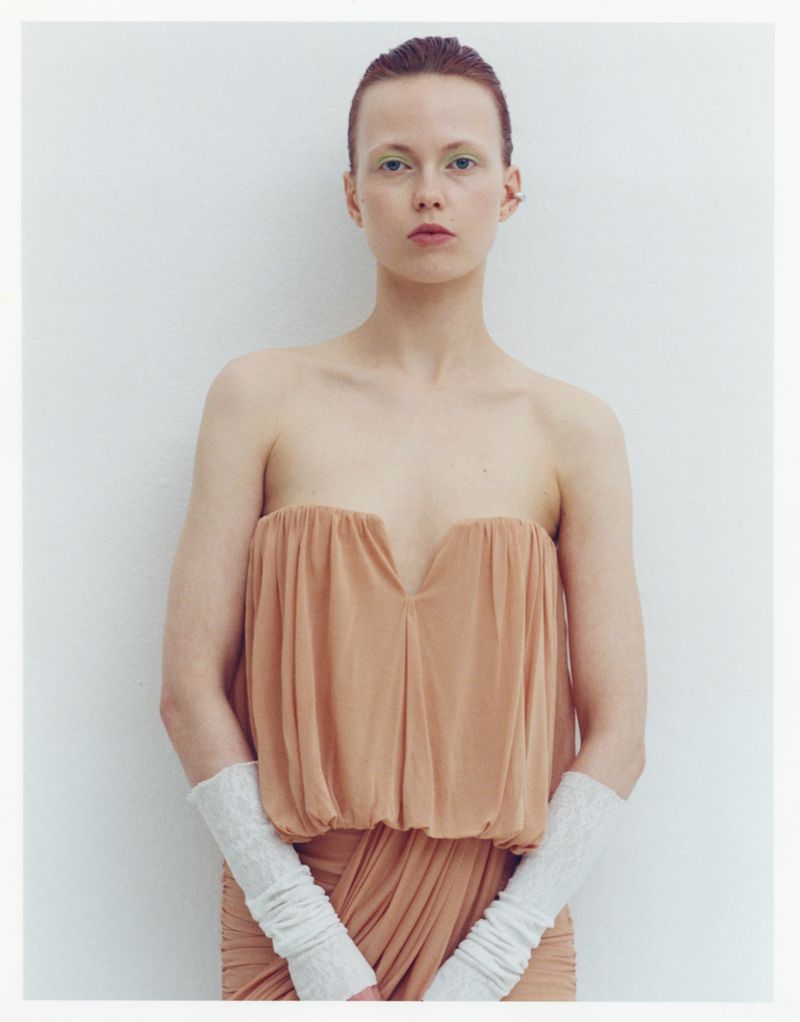 Woman in peach dress and knit gloves stands against a white wall in a studio portrait showcasing fashion and beauty aesthetic.