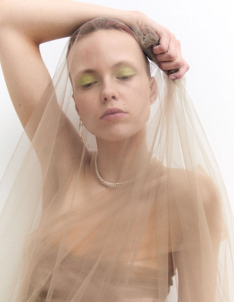 Portrait of a young adult woman with a sheer beige veil, wearing a necklace and green eyeshadow, set against a clean white backdrop.
