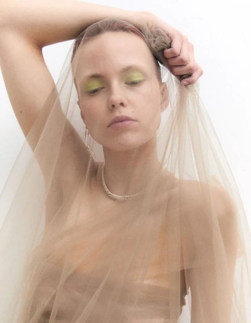 Portrait of a young adult woman with a sheer beige veil, wearing a necklace and green eyeshadow, set against a clean white backdrop.