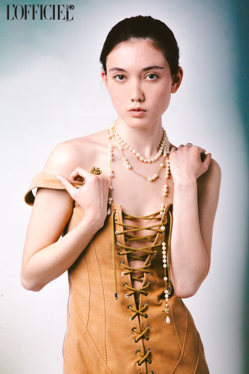 Portrait of a young woman wearing a beige dress with lace-up detail, layered pearl necklaces, and a gold ring, against a soft gradient backdrop.
