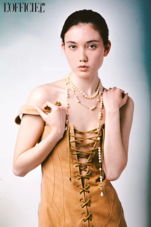 Portrait of a young woman wearing a beige dress with lace-up detail, layered pearl necklaces, and a gold ring, against a soft gradient backdrop.