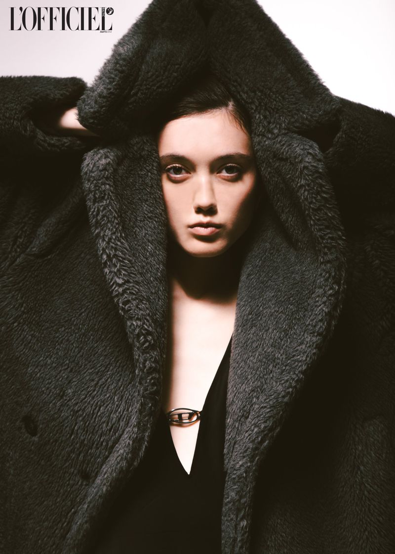 Fashion shot of a woman with dark hair wearing a large, dark grey faux fur coat and black top indoors against a white background.