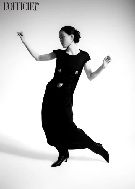Black and white studio shot of an Asian female model in an elegant black dress, high heel shoes, and minimalist makeup, posing expressively.