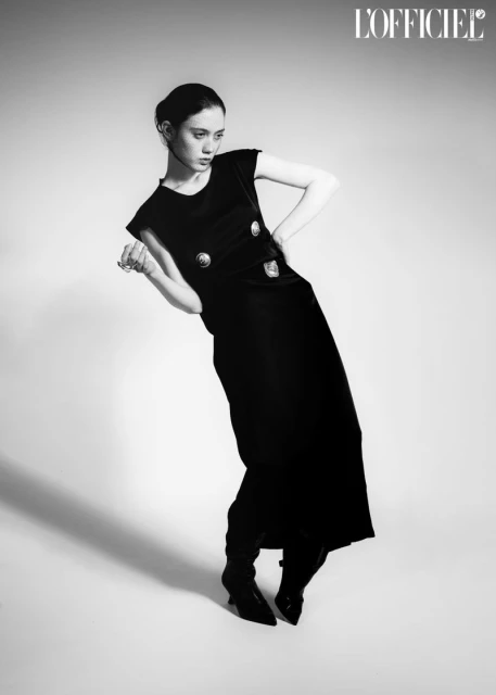 Black-and-white fashion portrait of a woman in an elegant black dress and boots, posing in a photo studio with soft, diffused lighting.