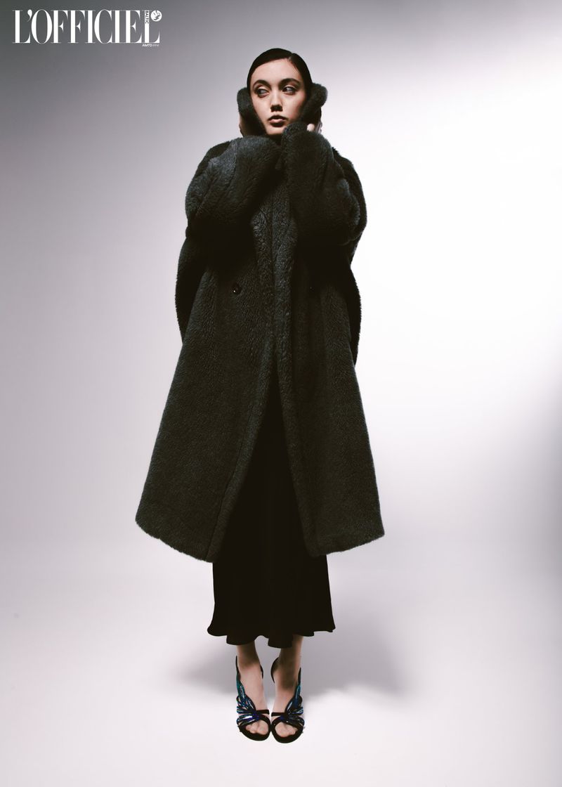 Full-length fashion shot of a female model wearing a long black coat, black dress, and sandals against a light gradient background.