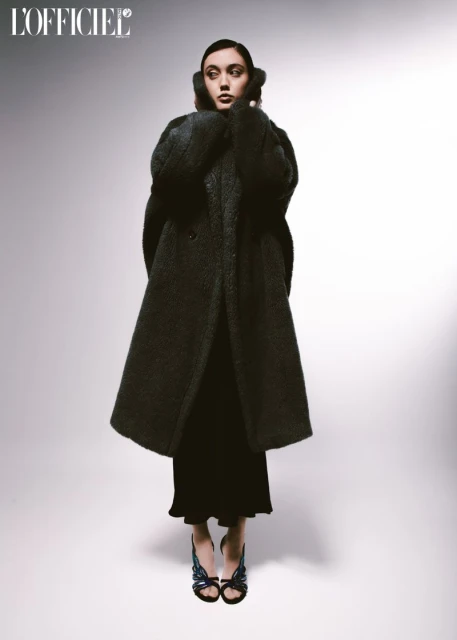 Full-length fashion shot of a female model wearing a long black coat, black dress, and sandals against a light gradient background.