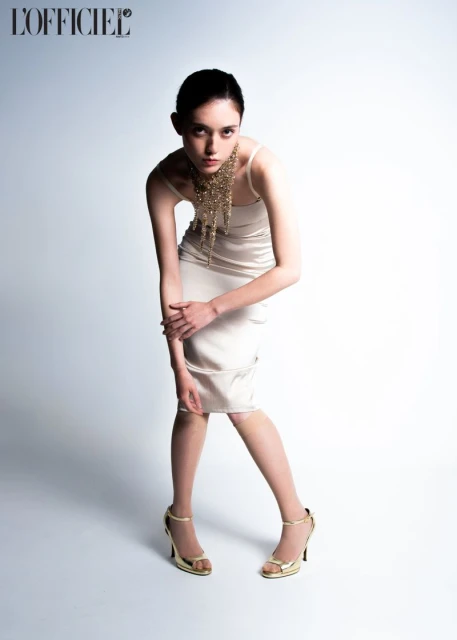 Fashion shot of a young adult woman in a light dress and gold heels, posing against a white background with a designer necklace.