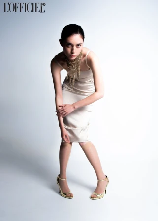 Fashion shot of a young adult woman in a light dress and gold heels, posing against a white background with a designer necklace.