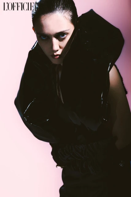 Fashion editorial shot of a young female model wearing a large, avant-garde shiny black vinyl dress against a gradient pink backdrop.