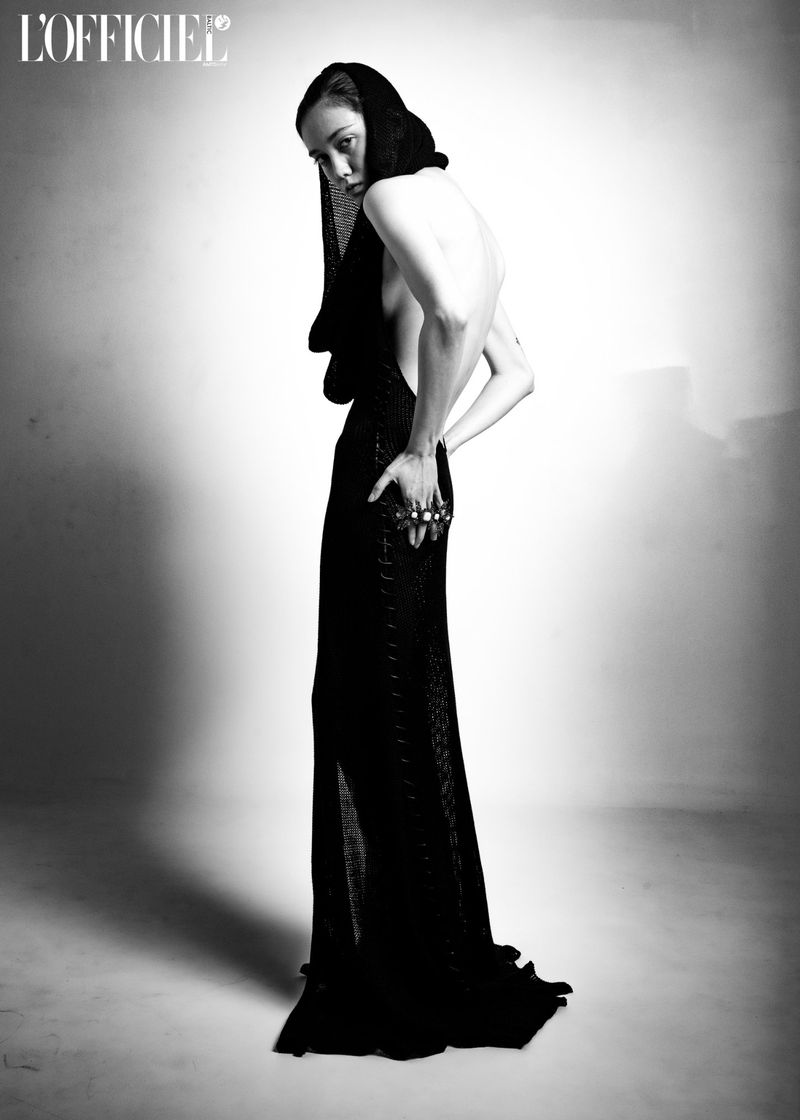 A slender woman poses in a black dress and head covering in a black and white studio fashion shot with soft lighting.
