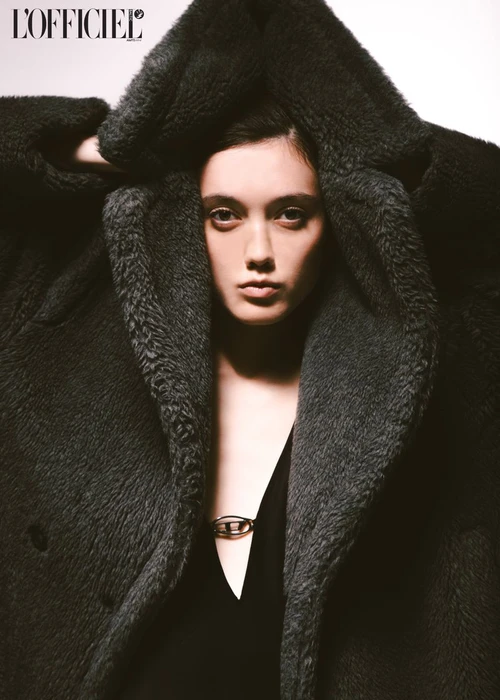 Model wearing a dark oversized furry coat with hood held up against a plain white background for L'Officiel Baltics Black Orchid editorial