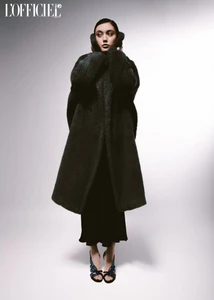 Model in a dark oversized coat and dark dress against a white gradient background for L'Officiel Baltics.