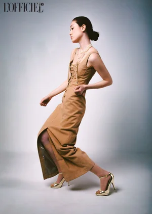 Model in a tan laced dress and gold heels posing against a grey studio backdrop for L'Officiel Baltics.