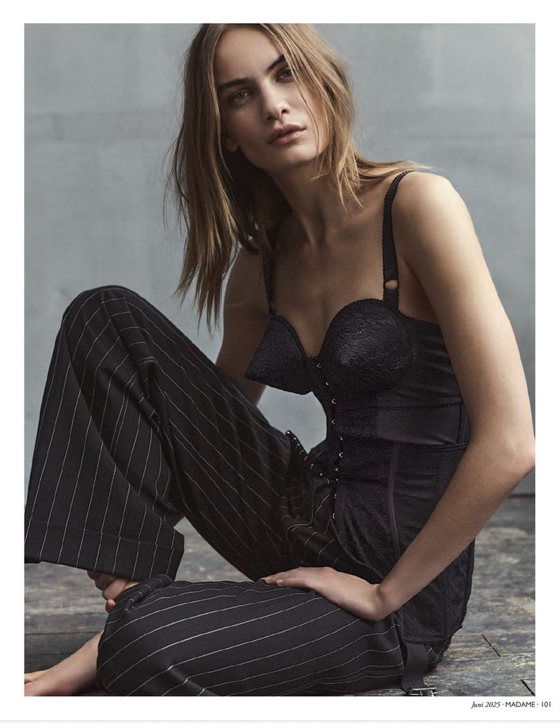 Elegant fashion shot of a slim woman with long hair wearing a black bustier top and striped trousers, seated indoors against a grey wall.