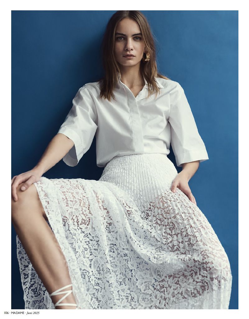 Fashion editorial shot of a woman in a white blouse and lace skirt against a dark blue background, posing in a relaxed sitting position.