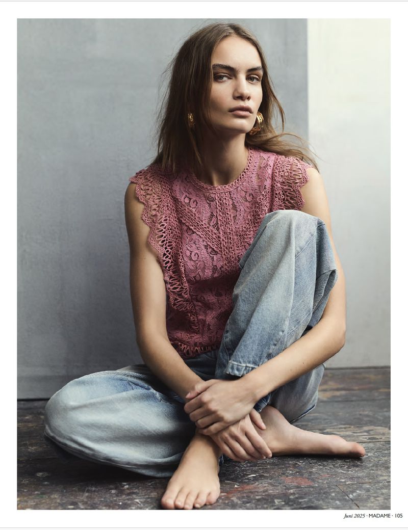 Portrait of a woman wearing a pink lace top and jeans, seated indoors. She has bare feet, brown hair, gold earrings and fair skin.