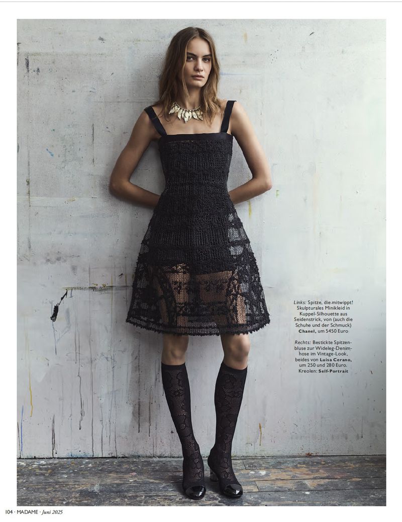 A woman in a black lace dress, knee-high patterned socks, and patent shoes poses against a textured wall in a fashion editorial shoot.