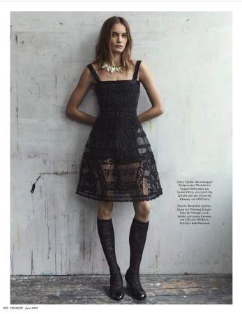 A woman in a black lace dress, knee-high patterned socks, and patent shoes poses against a textured wall in a fashion editorial shoot.