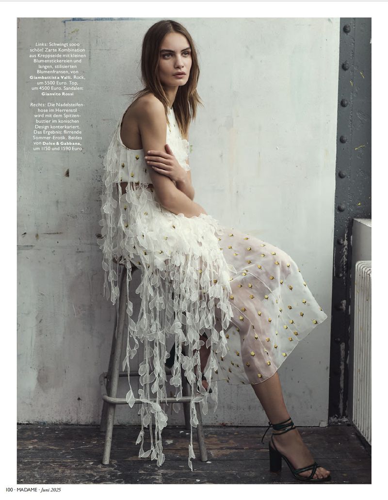 A woman in a white dress with decorative fringe poses while seated on a gray stool in a fashion-style medium indoor shot.